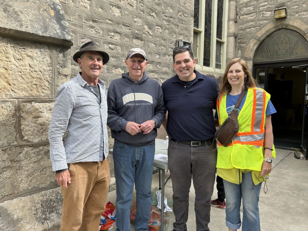Rodrigo is wearing the blue polo and gray pants. He is posing with a man wearing a hat (Mike Damon), a man wearing a baseball cap and a woman wearing a yellow safety vest (Claire Braden).